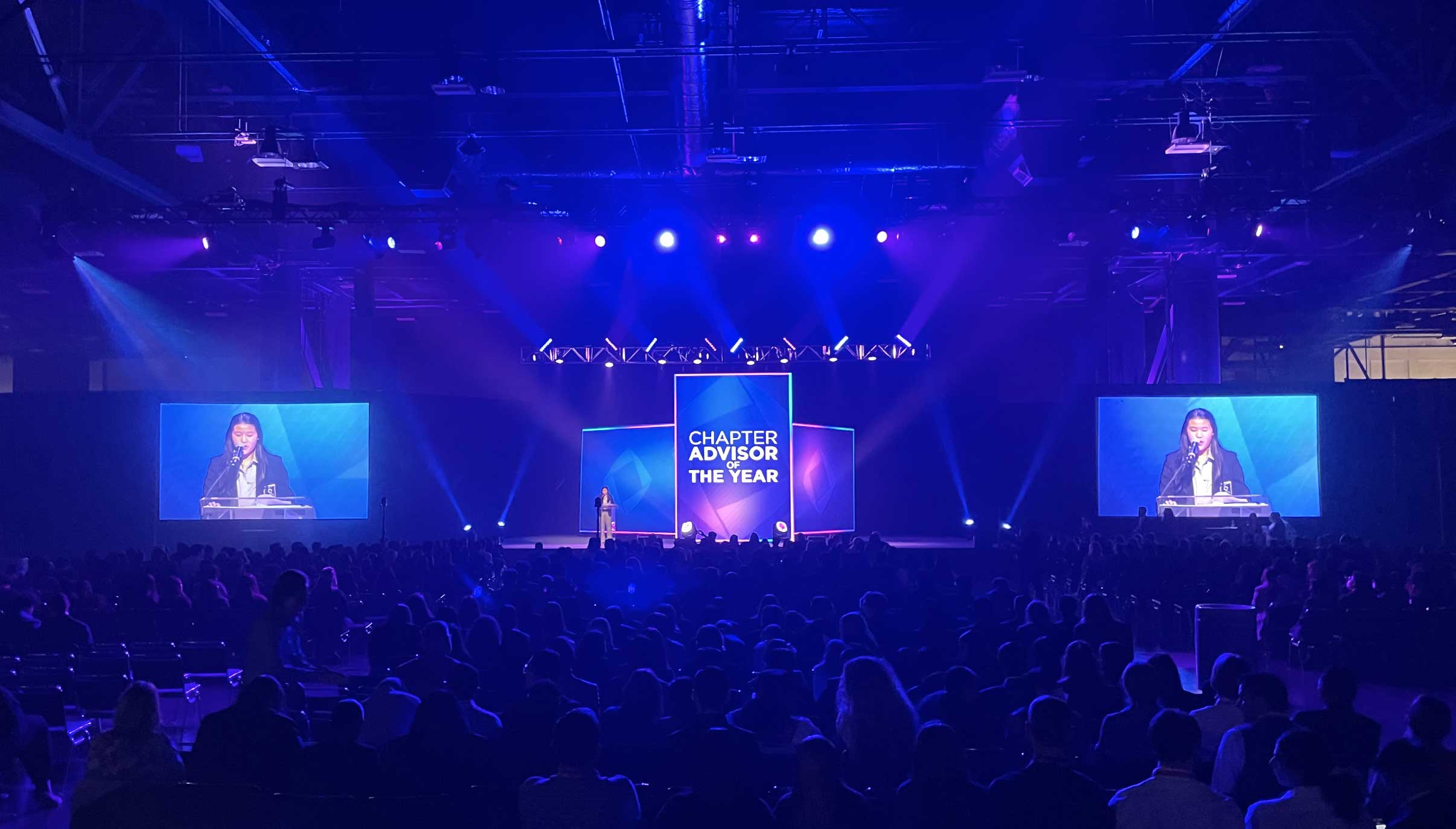 Full auditorium lit in blue with Chapter Advisor of the Year projected onstage.
